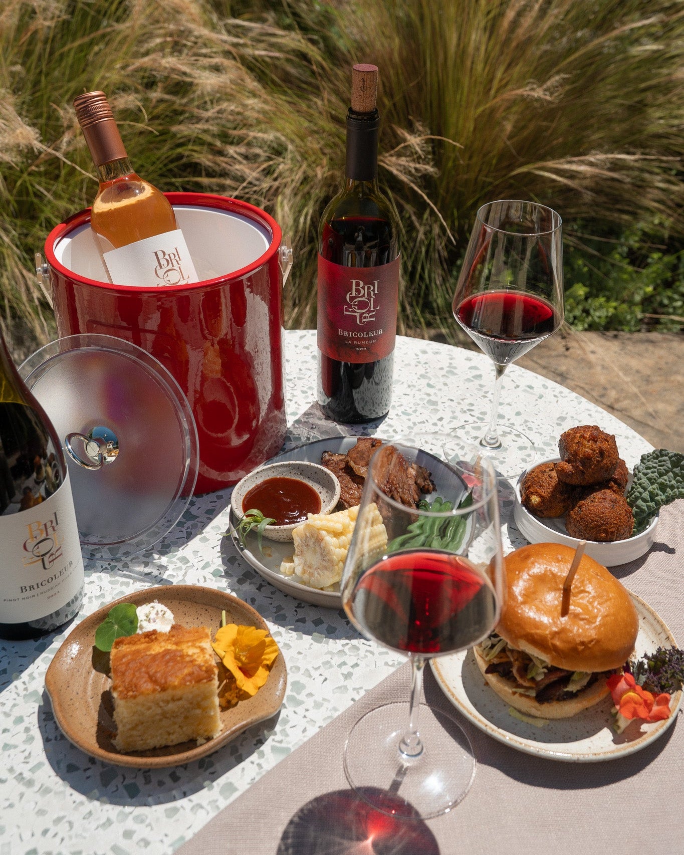 Outdoor dining setup with burgers, wine, and appetizers on a table with a natural background.