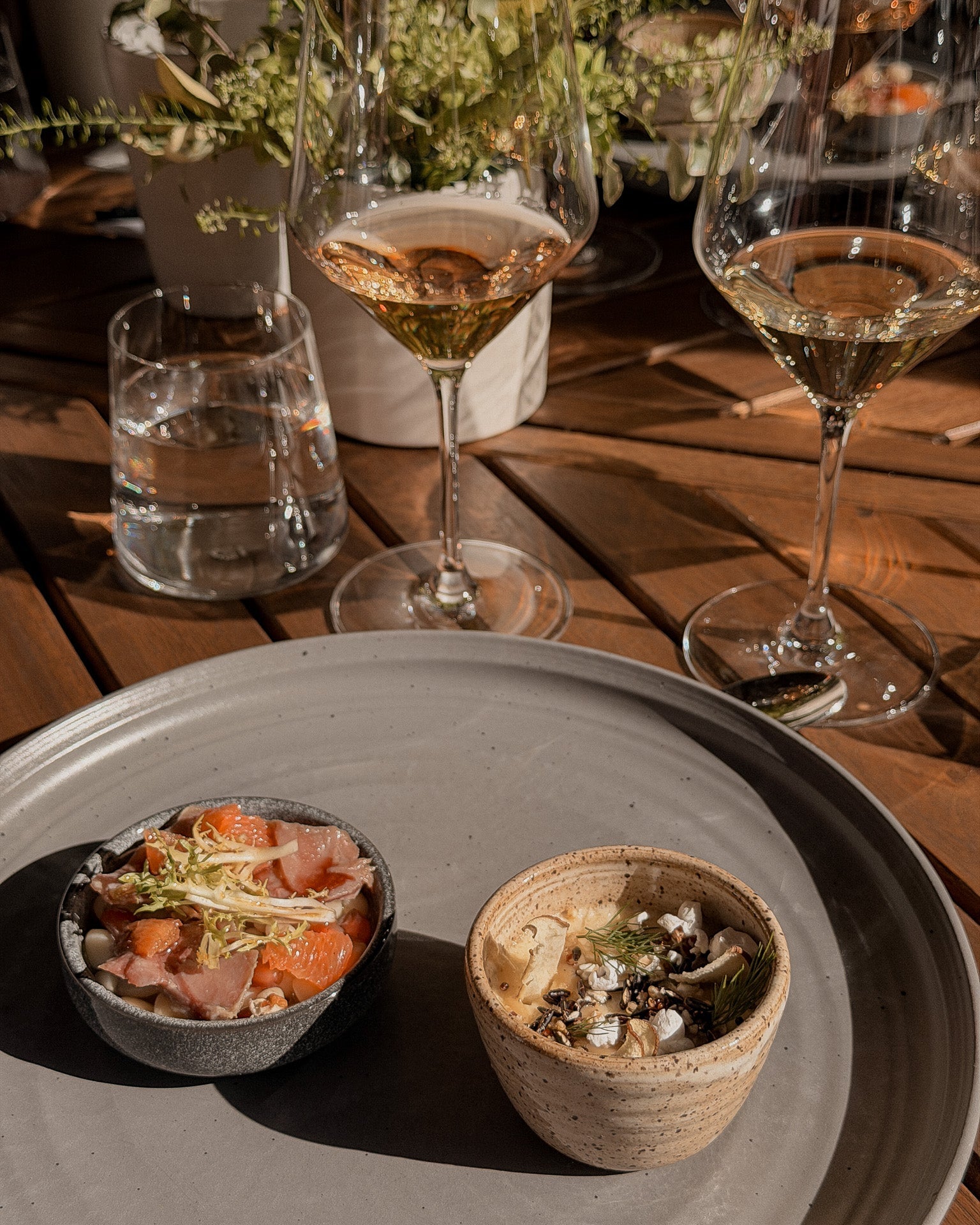 Two small bowls of food on a large plate with glasses of wine and water on a wooden table.